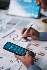 Fototapeta premium Young adult Black man holding smartphone with notifications visible while analyzing printed charts and graphs at desk, multitasking with pen in hand and digital device in workspace