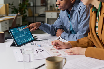 Young adult Black man and young adult Caucasian woman analyzing financial data on digital tablet, discussing charts and graphs, collaborating on business project at office desk