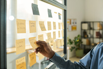Black man organizing sticky notes on glass wall, managing project tasks in modern office setting, hand reaching toward notes with to do list visible, focusing on workflow planning