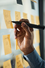 Black man hand writing on sticky notes attached to glass wall, planning or organizing tasks in office setting, visible wrist and part of sleeve suggesting young adult professional © pressmaster