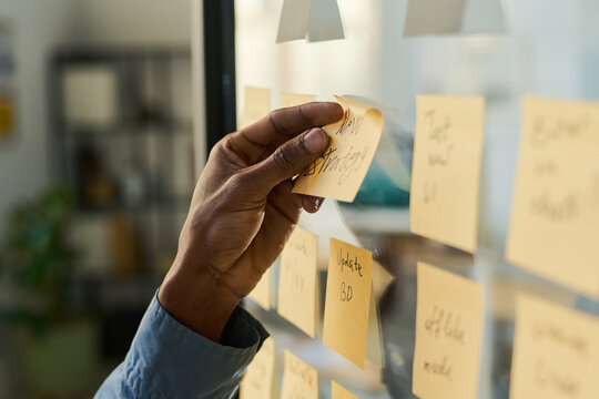 Black man organizing sticky notes on glass wall, hand placing note labeled new strategy among various reminders, business planning or brainstorming session in modern office setting