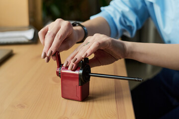 Young adult woman sharpening pencil using manual pencil sharpener on wooden desk, hands visible adjusting crank handle, wearing wristwatch, natural nails, close up