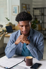 Young adult Black man sitting at desk contemplating, paperwork and coffee mug on table, smartphone nearby, office shelves and window in background