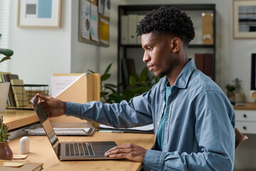 Young adult Black man sitting at desk using laptop, adjusting screen while working in modern office setting, focused expression, bookshelf and documents in background
