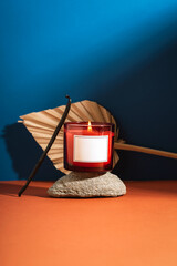 Red candle with flame placed on stone with dried palm leaf on blue and orange background. Aromatherapy and cozy home atmosphere concept. Mockup