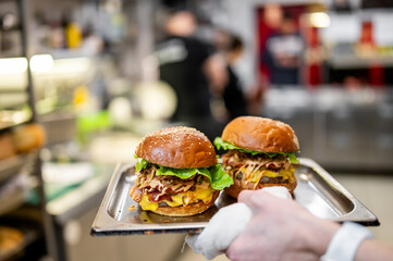 Two gourmet burgers on a metal serving tray in a commercial kitchen setting. Hand holding tray with...