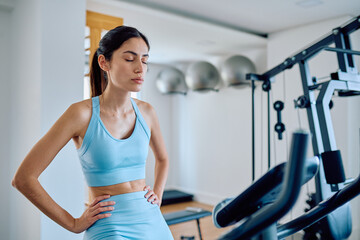 Fototapeta premium Athletic woman standing with hands on her hips, taking a deep breath and resting between sets in the gym