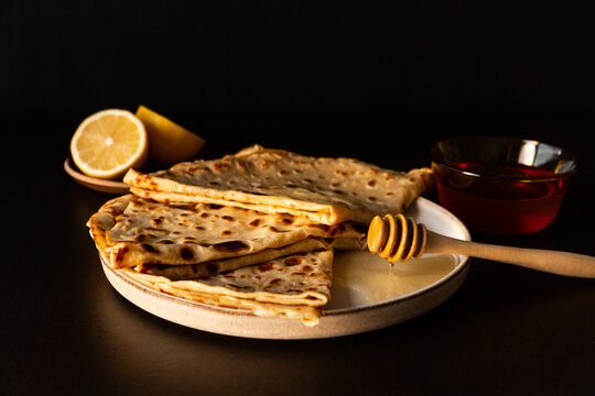Gros plan sur des cr&ecirc;pes sucr&eacute;es pour la chandeleur ou mardi gras, pr&eacute;sent&eacute;es sur une assiette avec du miel et un citron coup&eacute;, couleurs gourmandes, vue de face sur un fond brun fonc&eacute;