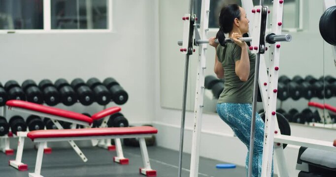 A middle-aged woman building leg muscles while squatting Smith machine in gym. Woman builds leg muscles with focused squats on the Smith machine in the gym. Slow motion 4k footage.