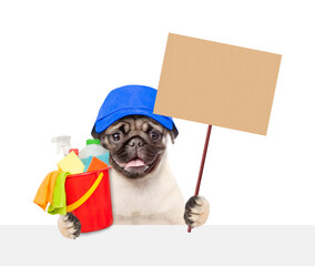 Cleaning concept. Funny Pug puppy wearing blue cap holds blank placard and bucket with  washing fluids, rags and spray in paws above blank white banner. isolated on white background