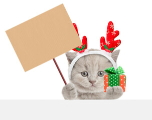 Funny tiny kitten dressed like santa claus reindeer  Rudolf holding blank placard anf gift box in it paw and looking above blank white banner. isolated on white background