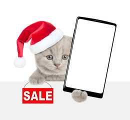 Cute kitten wearing red christmas hat holds smartphone with white blank screen in it paw looks above blank white banner. and showing signboard with labeled "sale". Empty spsce. Isolated on white