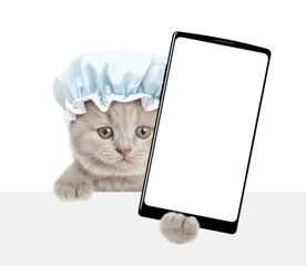 Funny tiny kitten wearing shower cap holds big smartphone with white blank screen in it paw above blank white banner. isolated on white background