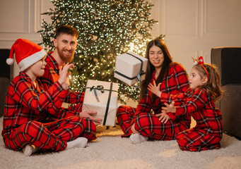 appy young family in matching red plaid pajamas having fun and tossing a gift box while sitting on the floor in front of a glowing Christmas tree at home.