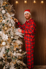 Happy man in red plaid pajamas and Santa hat decorating a Christmas tree with gold ornaments at home.