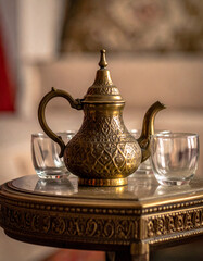 A close-up of a small, decorative, hammered brass tea pot resting on a low, ornate table next to three small, empty, clear glasses.