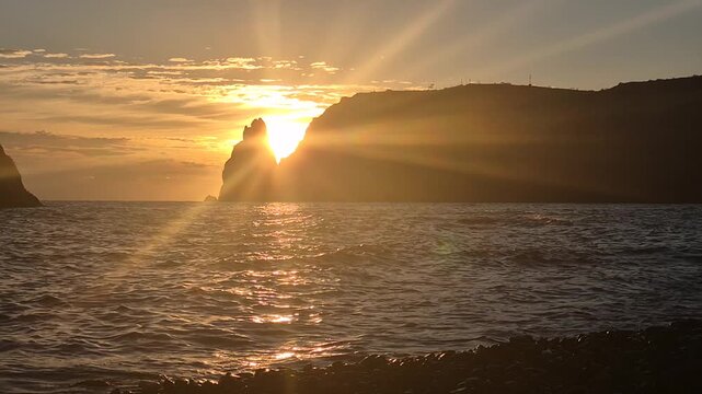 Sunset ocean cliff. Stunning golden hour sun rays burst behind the towering rock formation over the sparkling sea. Vertical video.