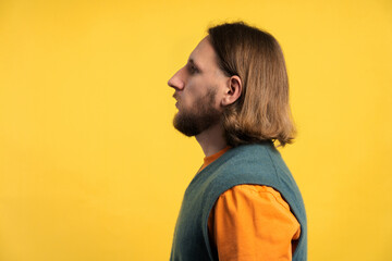 Young man with beard and long hair standing in profile against yellow background