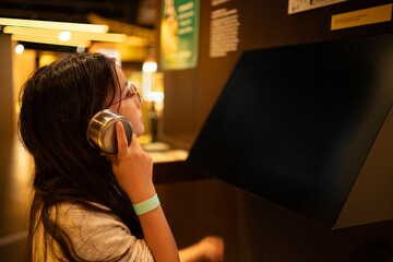 Teenage girl in glasses holding headphone to ear while looking at museum exhibit, education and curiosity concept