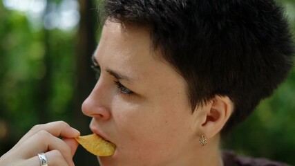 Close-up of a woman with short dark hair eating crispy potato chips while relaxing outdoors in a sunny green park. 4k horizontal footage. The concept of delicious fatty and unhealthy food