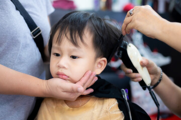 Close-up of a stylist using a hair clipper to trim a young Asian boy's hair at a salon....