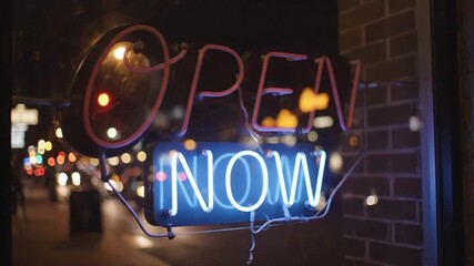 Vibrant red and blue neon OPEN NOW sign glows in a shop window at night, with city street lights blurred in the background, inviting customers.