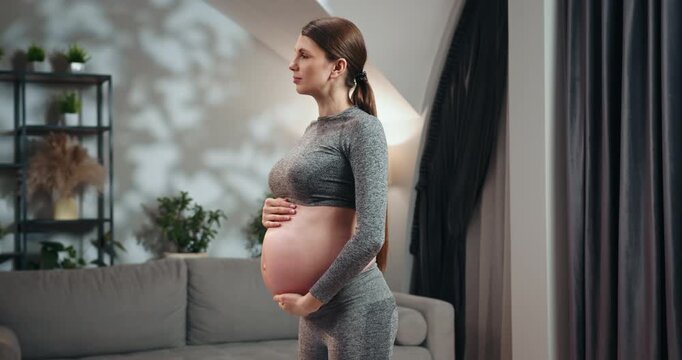 Pregnant woman in athletic wear cradles her belly, standing in a softly lit room with home decor elements visible. Pregnant Woman Holding Belly