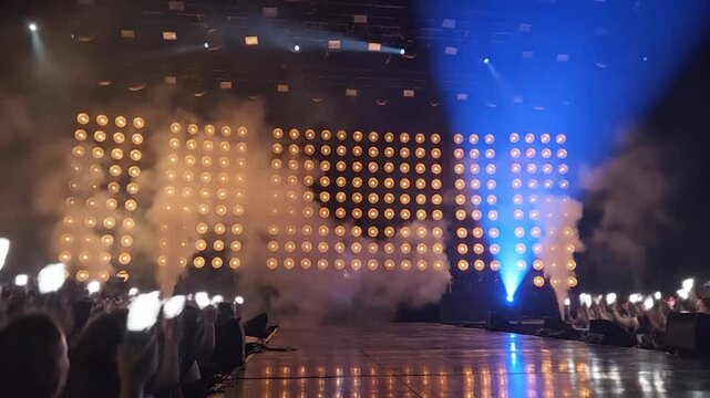 Empty concert stage with a massive LED wall, dramatic smoke, and pyrotechnics, as an excited crowd holds up phones under colorful spotlights.