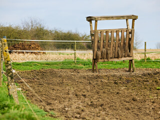 Rustic wooden hay feeder stands in a muddy paddock, framed by a simple fence and green grass. The rural landscape with trees in the background suggests a farm or countryside setting.