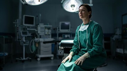 Female surgeon in green scrubs and face mask hanging from ear, sitting on a stool in a dimly lit operating room, looking down with a sad and stressed expression after a difficult surgery
