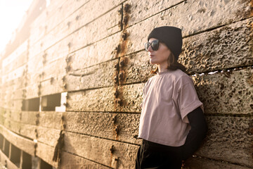 woman in a pink T shirt, black beanie, and sunglasses rests against a textured, sunlit wall