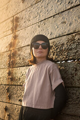 woman in a pink T shirt, black beanie, and sunglasses rests against a textured, sunlit wall