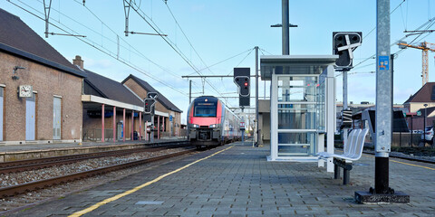 A modern train pulls into a station platform with tracks, overhead lines, and a brick station building under a clear blue sky.