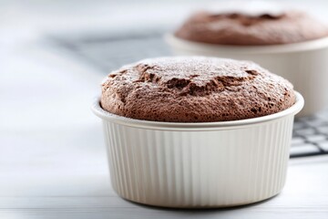 Chocolate souffl&eacute; in white ramekin stands on white table, sprinkled with powdered sugar, second souffl&eacute; in background.