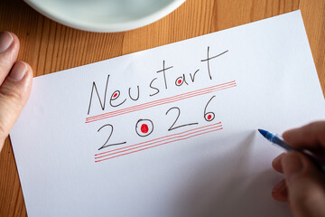 Nahaufnahme einer Hand, die Neustart 2026 auf ein wei&szlig;es Blatt Papier schreibt. Symbolbild f&uuml;r neue Anf&auml;nge und Jahresplanung auf Holzgrund