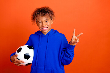 Young boy in blue hoodie holds soccer ball and shows peace sign against bright orange background