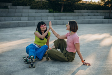 Glad women give high five and hug sitting on asphalt square after riding roller skating in park