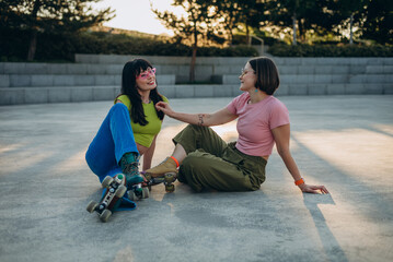 Pretty women friends with quad skates rest sitting on asphalt square
