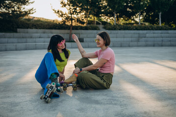 Young girl friends rest sitting on asphalt square after riding at sunset
