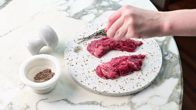 Preparing meat on a marble countertop, seasoning with spices in a kitchen setting during daytime