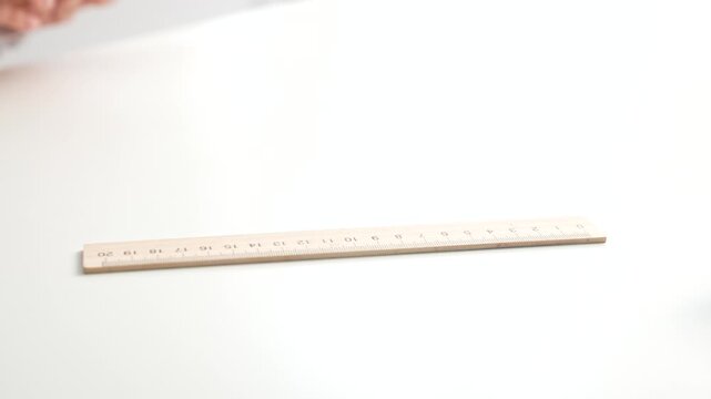 A hand places a wooden ruler on a white tabletop indoors.