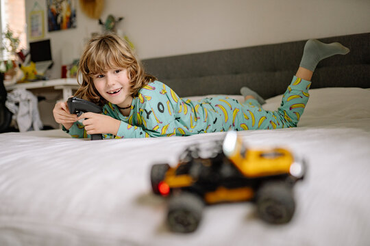 Boy plays with toy car in pajamas on bed in his bedroom during the day