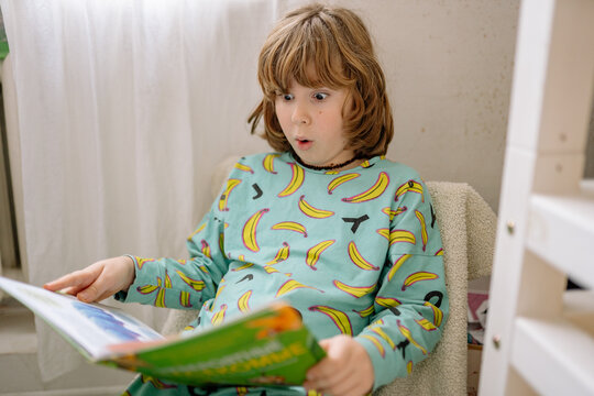 Boy reads a book in his bedroom while sitting on a chair in pajamas