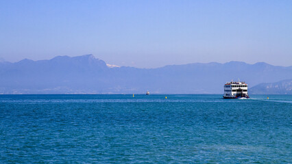 A passenger ferry sailing across the calm blue waters of Lake Garda, against the backdrop of a...