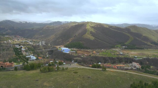 Aerial drone video of jermuk city in mountain valley of armenia, showing urban neighborhoods, green parks and dramatic hills, bridges and roads with calm cinematic movement in daylight soft tone