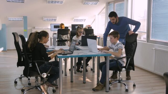 Teachers explaining to students working on laptops in modern, bright classroom during STEM lesson