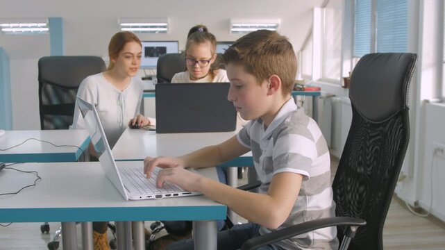 Students using laptops in modern classroom with teacher helping girl in background