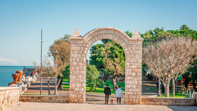A free-standing, pointed limestone archway marks the entrance to a lush seaside park in Trani. Two people walk a dog toward green trees and a playground, with the blue Adriatic Sea visible to the left