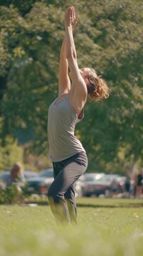 Woman practicing yoga outdoors in a green park, standing in an upward stretch pose with arms raised toward the sky.