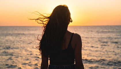 Fototapeta premium Back view of woman looking at ocean sunset, hair flowing in the sea breeze, golden hour glow